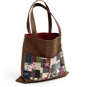 Op de afbeelding: Bruine tote bag met een patchwork zak. De tas heeft een rode binnenkant en bruine handvatten. De patchwork zak heeft verschillende stofpatronen in tinten blauw, groen, roze en wit. De tas is gemaakt van een stevig materiaal.