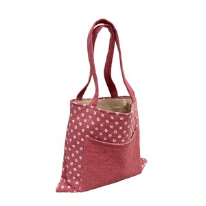 Könnte beinhalten: Eine rote Tragetasche mit weißen Blumenmustern. Die Tasche hat eine große Vordertasche und zwei Henkel. Das Innenfutter ist hellbeige. Die Tasche ist aus einem strukturierten Stoff gefertigt.