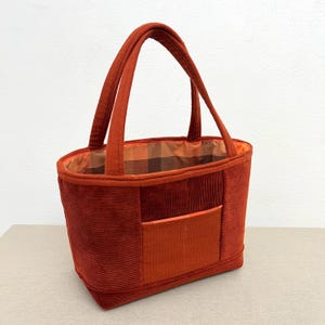Può includere: Una borsa tote in velluto a coste color ruggine con due manici robusti e una tasca frontale rettangolare. L'interno è foderato con un tessuto a motivo scozzese marrone e arancione, che contrasta con la superficie esterna testurizzata.