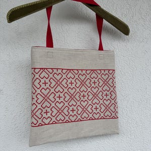 Pode incluir: Uma bolsa tote bege com uma larga faixa horizontal de padrões de corações em ponto cruz vermelho. A bolsa tem duas alças de tecido vermelho e está pendurada num cabide de veludo verde. O design consiste em formas de coração repetidas.