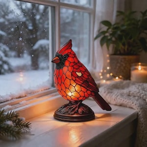 May include: A stained glass cardinal lamp in shades of red, orange, and black sits on a windowsill. The bird-shaped lamp has a detailed mosaic pattern and is perched on a bronze-colored base. The background shows a snowy outdoor scene.