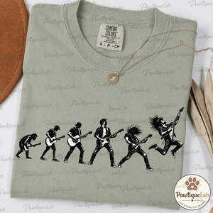 Puede incluir: Una camiseta verde salvia con un estampado gráfico negro que muestra la evolución de un guitarrista, desde una figura encorvada hasta una estrella de rock saltando mientras toca una guitarra eléctrica. La camiseta tiene cuello redondo y etiqueta Comfort Colors.