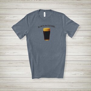 May include: A slate blue t-shirt featuring the text "WHEN IN DOUBT" above a graphic of a pint of dark beer. The beer has a golden head and a dark brown body. The shirt is made of a soft material.