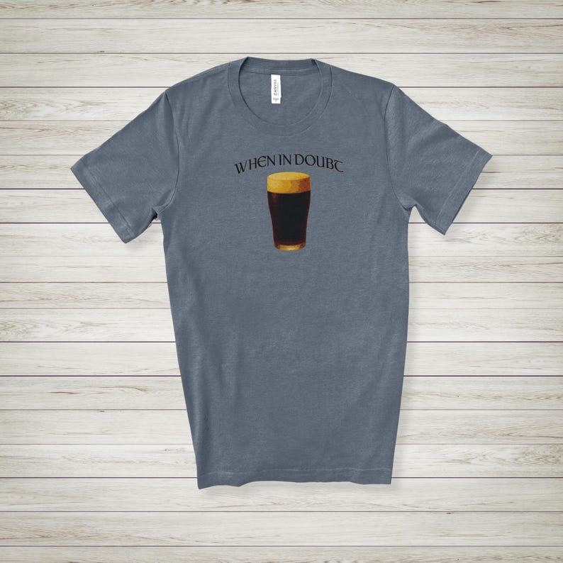 May include: A slate blue t-shirt featuring the text "WHEN IN DOUBT" above a graphic of a pint of dark beer. The beer has a golden head and a dark brown body. The shirt is made of a soft material.