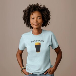 May include: A light blue t-shirt with the text "WHEN IN DOUBC" above a graphic of a pint of dark beer. The shirt is short-sleeved and has a crew neck. The person is wearing a gold bracelet.