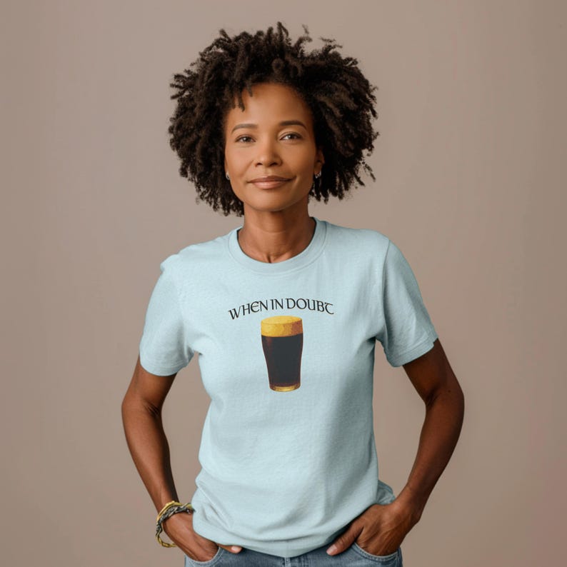 May include: A light blue t-shirt with the text "WHEN IN DOUBC" above a graphic of a pint of dark beer. The shirt is short-sleeved and has a crew neck. The person is wearing a gold bracelet.