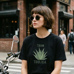 Puede incluir: Una camiseta negra con las palabras "TEAM TEQUILA" arriba y abajo de una ilustración de agave verde. La persona de la imagen lleva gafas de sol y está cerca de una motocicleta.