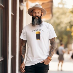 May include: White t-shirt with the text "WHEN IN DOUBC" above a graphic of a pint of dark beer. The shirt is being worn by a person with a long beard and a hat.