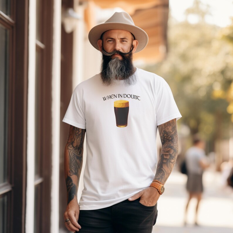 May include: White t-shirt with the text "WHEN IN DOUBC" above a graphic of a pint of dark beer. The shirt is being worn by a person with a long beard and a hat.