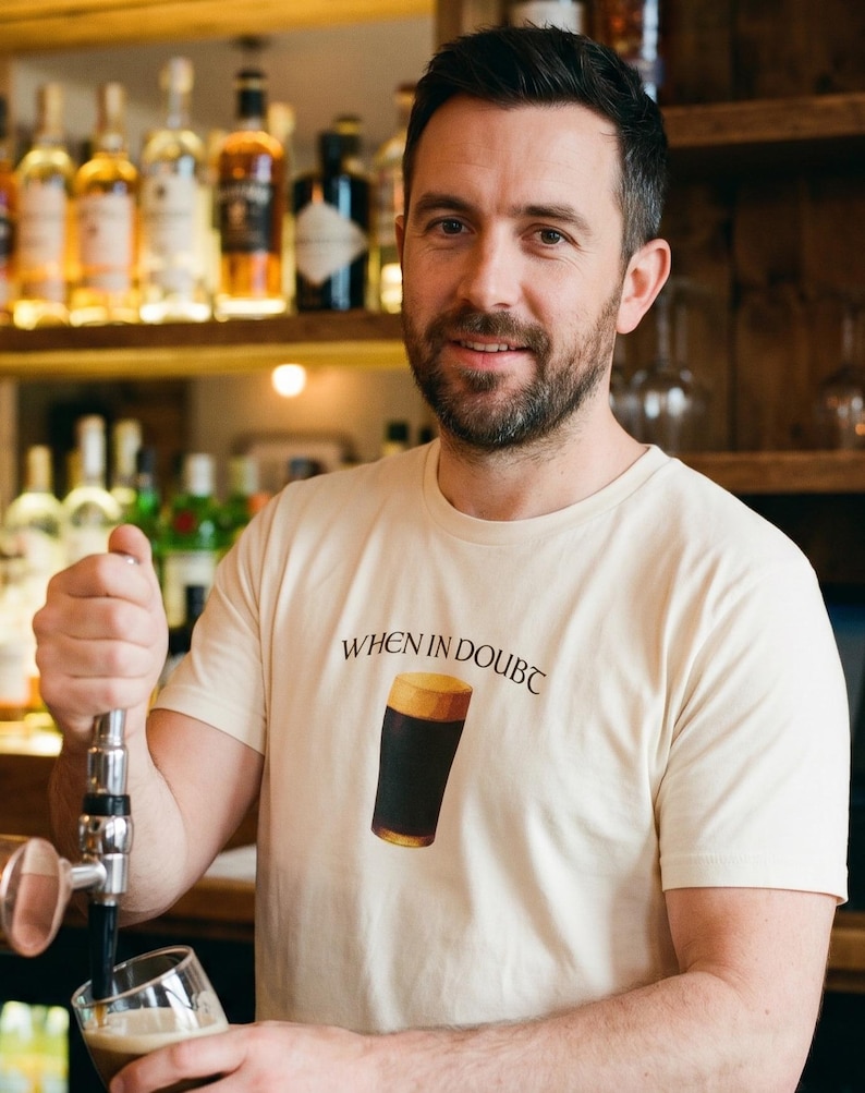 May include: A man wearing a cream-colored t-shirt with the text "WHEN IN DOUBT" above a graphic of a pint of dark beer. The man is pouring beer from a tap into a glass. Shelves of liquor bottles are in the background.