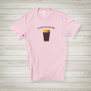 May include: A light pink t-shirt with the text "WHEN IN DOUBC" above a graphic of a pint of dark beer. The beer has a dark brown body and a tan head. The shirt is made of a soft material.