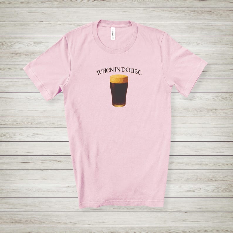 May include: A light pink t-shirt with the text "WHEN IN DOUBC" above a graphic of a pint of dark beer. The beer has a dark brown body and a tan head. The shirt is made of a soft material.