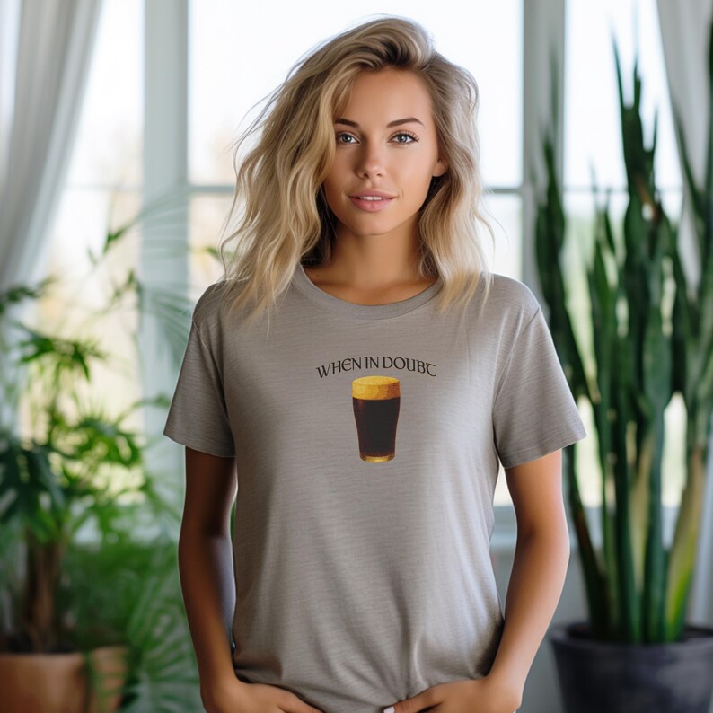 May include: A light gray t-shirt featuring the text "WHEN IN DOUBC" above a graphic of a pint of dark beer with a golden head. The shirt is a crew neck style.
