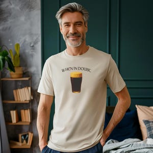 May include: Beige t-shirt with the text "WHEN IN DOUBC" above a graphic of a pint of dark beer. The shirt is short-sleeved and worn by a person with gray hair and a beard.