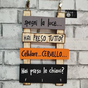 Può includere: Un portachiavi in legno con quattro assi orizzontali di vari colori e testo. Il testo include frasi come "Spegni la luce" e "Hai preso tutto?" Il portachiavi ha tre ganci ed è sospeso da una corda.