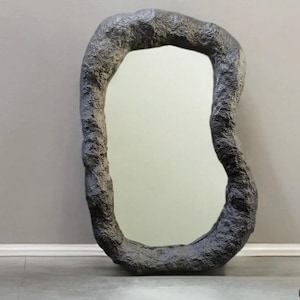 Può includere: Uno specchio da terra caratterizzato da una cornice dalla forma organica irregolare con una finitura scura, ruvida e simile alla pietra. La superficie in vetro riflettente è racchiusa in questo bordo spesso che imita la roccia.