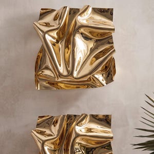 May include: Two square wall art pieces featuring a highly reflective, polished gold finish. Each piece has a deeply creased, folded, and undulating surface texture that creates complex light reflections and shadows across the metallic material.