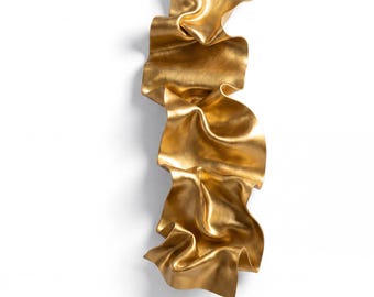 May include: A vertical wall sculpture featuring a continuous, flowing ribbon-like form with a metallic gold finish. The object displays deep, undulating folds and curves that create a sense of movement and texture across its elongated surface.