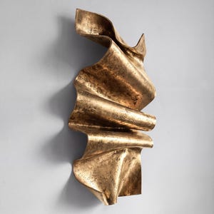 May include: A vertical wall sculpture featuring a metallic gold finish with a textured, weathered surface. The object has a fluid, undulating form with deep folds and curves that create a sense of movement, resembling draped fabric or ribbon.