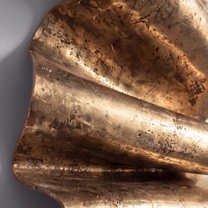 May include: A metallic wall sculpture featuring a series of undulating, wave-like folds. The surface has a burnished gold finish with a distressed, textured appearance characterised by dark, mottled markings and fine scratches across the metal.
