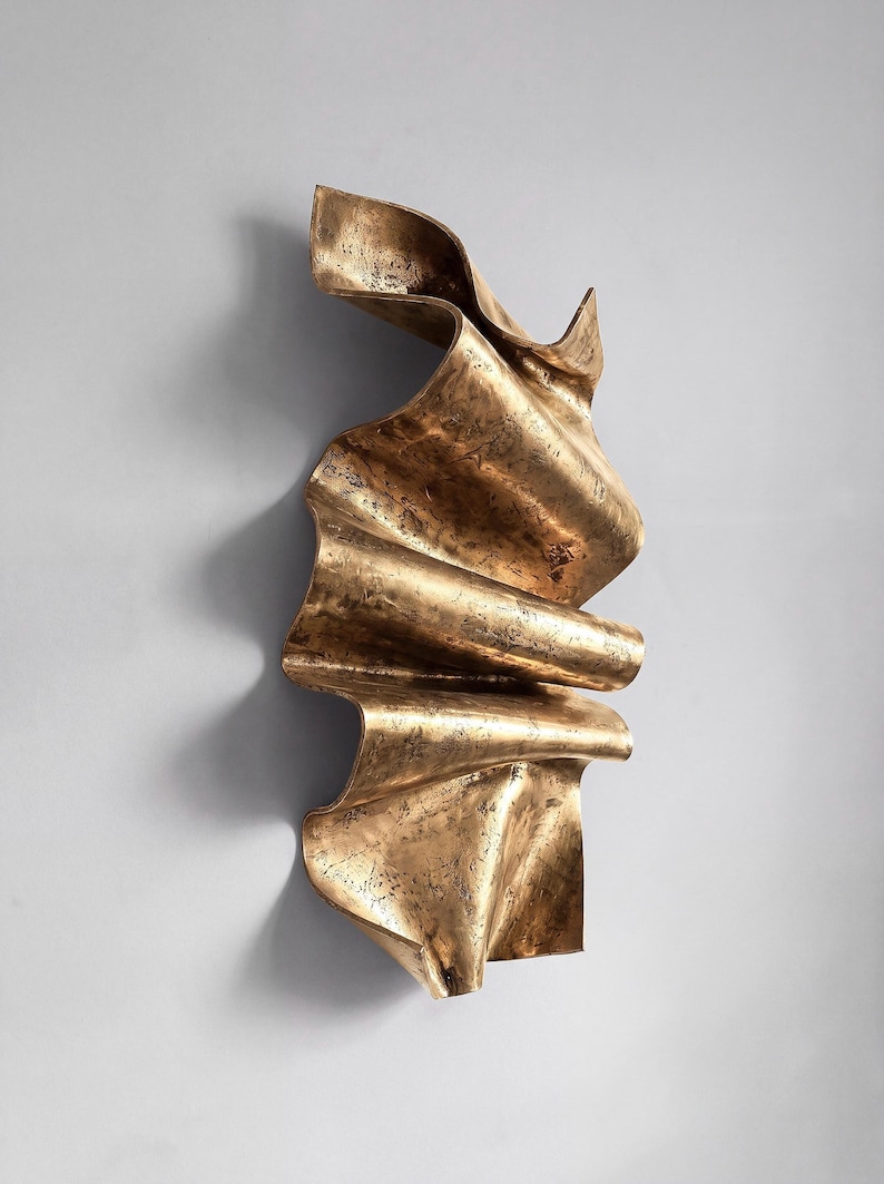 May include: A vertical wall sculpture featuring a metallic bronze finish with a textured, weathered surface. The object has a fluid, undulating form with deep folds and curves that create a sense of movement, resembling a piece of draped fabric.