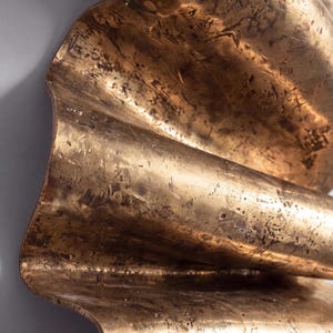 May include: A metallic wall sculpture featuring a series of undulating, wave-like folds. The surface has a burnished gold finish with a distressed, textured appearance characterised by dark, mottled markings and fine scratches across the metal.