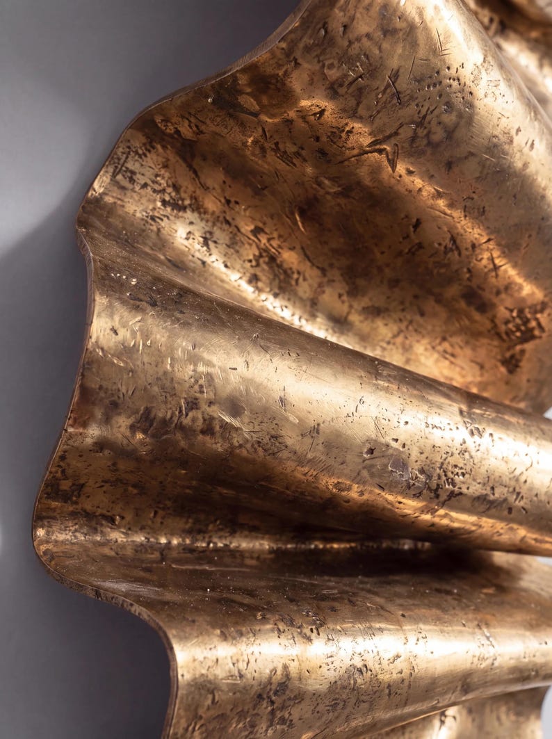 May include: A metallic wall sculpture featuring a series of undulating, wave-like folds. The surface has a burnished gold finish with a distressed, textured appearance characterised by dark, mottled markings and fine scratches across the metal.