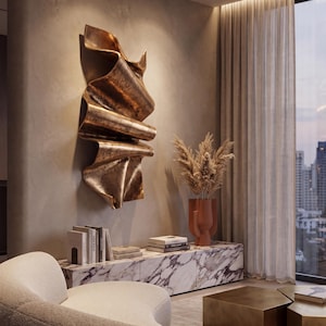 May include: A large, vertical, bronze-coloured wall sculpture with a folded, ribbon-like texture mounted on a beige wall above a white marble console table that holds several books and a terracotta-coloured vase filled with dried pampas grass.