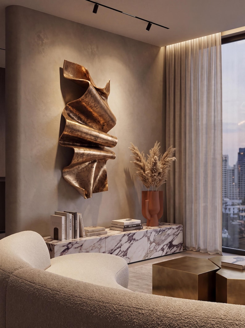 May include: A large, vertical, bronze-coloured wall sculpture with a folded, ribbon-like texture mounted on a beige wall above a white marble console table that holds several books and a terracotta-coloured vase filled with dried pampas grass.