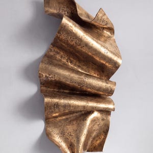 May include: A vertical wall sculpture made of bronze-toned metal with a rippled, wave-like form. The surface features a textured, mottled finish with dark brown and metallic gold highlights, creating a sense of movement and depth in the folds.