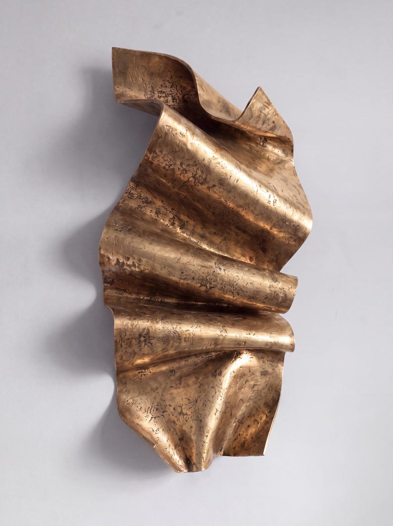 May include: A vertical wall sculpture made of bronze-toned metal with a rippled, wave-like form. The surface features a textured, mottled finish with dark brown and metallic gold highlights, creating a sense of movement and depth in the folds.
