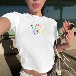 May include: White crop top with a colorful embroidered design of four cartoon characters. The design features green, red, blue, and yellow figures. The shirt is short-sleeved and appears to be made of a soft material.