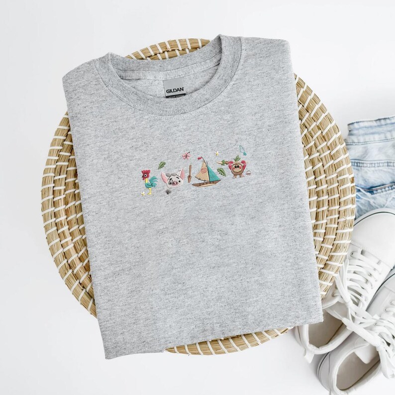 May include: A heather gray crewneck sweatshirt featuring a colorful embroidered design of cartoon characters, a sailboat, and musical notes. The sweatshirt is made of a soft material and is displayed on a woven basket.