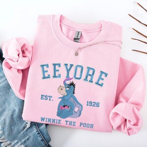 May include: A light pink sweatshirt featuring the word "Eeyore" in blue lettering above an illustration of Eeyore from Winnie the Pooh. The sweatshirt also includes the text "EST. 1926" and "WINNIE THE POOH". A silver necklace with a sun pendant is on the sweatshirt.