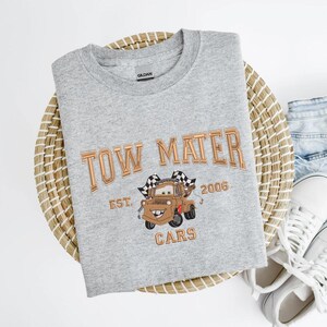 Puede incluir: Sudadera gris claro con cuello redondo y texto bordado en cobre "TOW MATER". Debajo, una imagen bordada de Mate de Cars, con el texto "EST. 2006 CARS" debajo.