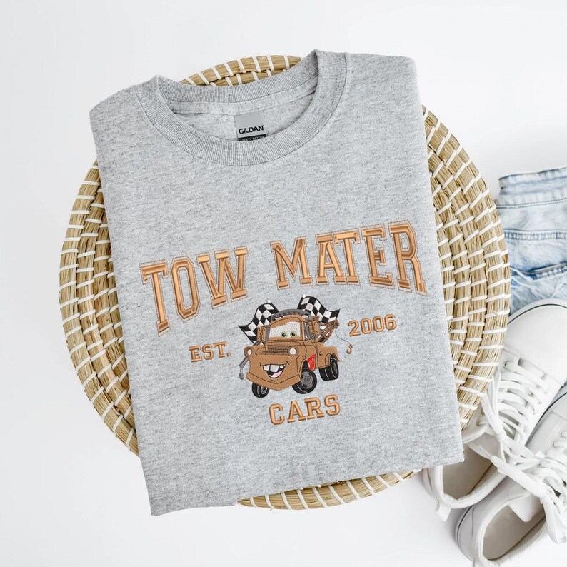 Puede incluir: Sudadera gris claro con cuello redondo y texto bordado en cobre "TOW MATER". Debajo, una imagen bordada de Mate de Cars, con el texto "EST. 2006 CARS" debajo.