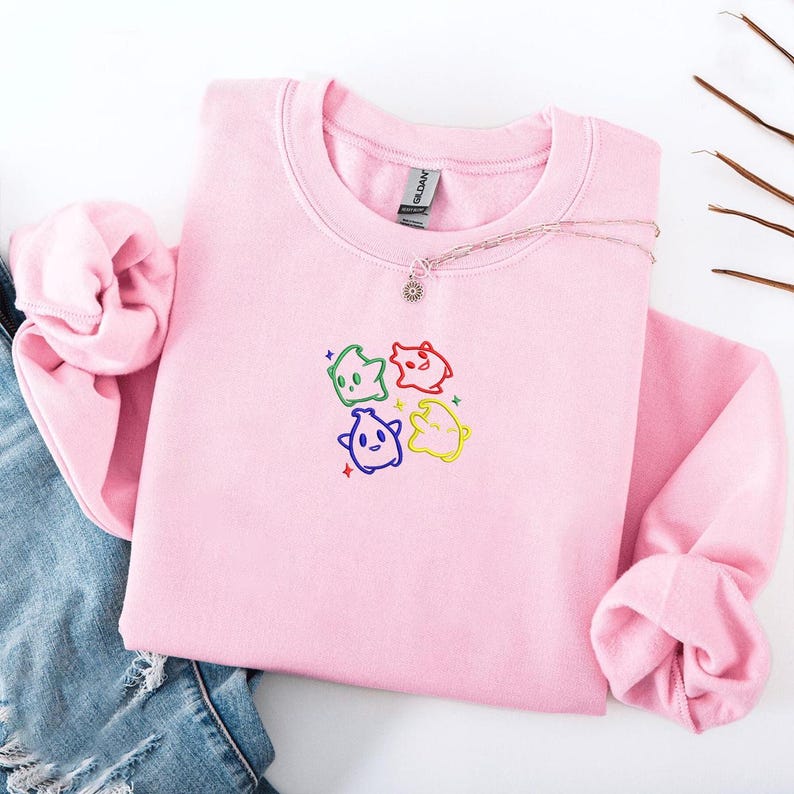 May include: A light pink sweatshirt with a crew neck. The front features an embroidered design of four colorful, cartoonish figures. A silver necklace with a small pendant rests on the sweatshirt. The sweatshirt is folded on a white surface.