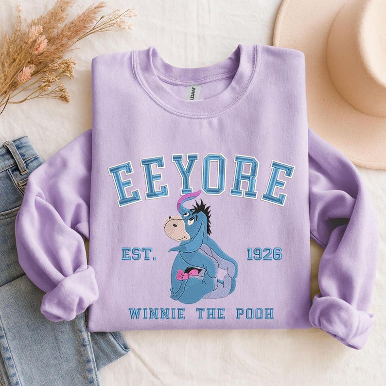 May include: A lilac-colored sweatshirt featuring the word "Eeyore" in a collegiate font above an illustration of Eeyore from Winnie the Pooh. The sweatshirt also includes the text "EST. 1926" and "WINNIE THE POOH".