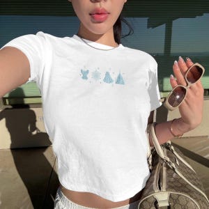May include: White crew neck t-shirt with a light blue embroidered design featuring a bird, snowflake, tent, and castle. The shirt is a cropped style. The person is holding sunglasses and a patterned handbag.