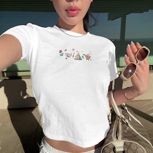 May include: White crop top with a colorful embroidered design. The design includes a rooster, a pig, a sailboat, and a bear. The shirt is short-sleeved and has a crew neck.