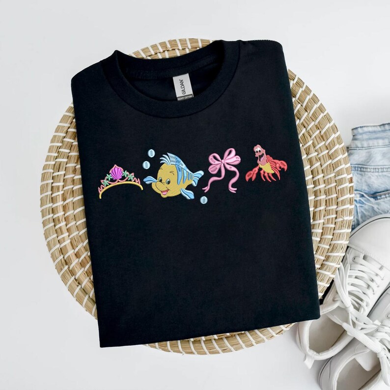 May include: Black t-shirt with embroidered cartoon characters from a popular animated film. The design includes a gold tiara, a yellow and blue fish, a pink bow, and a red crab. The shirt is displayed on a woven basket.