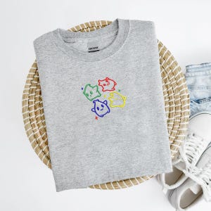 May include: A heather gray crew neck sweatshirt featuring an embroidered design of four colorful cartoon ghosts. The ghosts are outlined in different colors: green, red, blue, and yellow, with matching star accents. The sweatshirt is displayed on a woven basket.