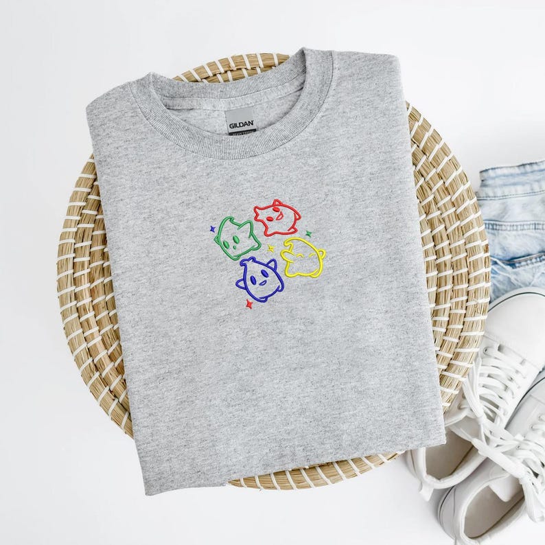 May include: A heather gray crew neck sweatshirt featuring an embroidered design of four colorful cartoon ghosts. The ghosts are outlined in different colors: green, red, blue, and yellow, with matching star accents. The sweatshirt is displayed on a woven basket.