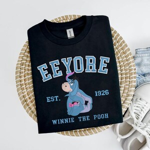 May include: Black t-shirt featuring a graphic of Eeyore from Winnie the Pooh. The design includes the name "Eeyore" in blue and white lettering, the year "1926", and the phrase "Winnie the Pooh". The shirt is displayed on a woven basket.