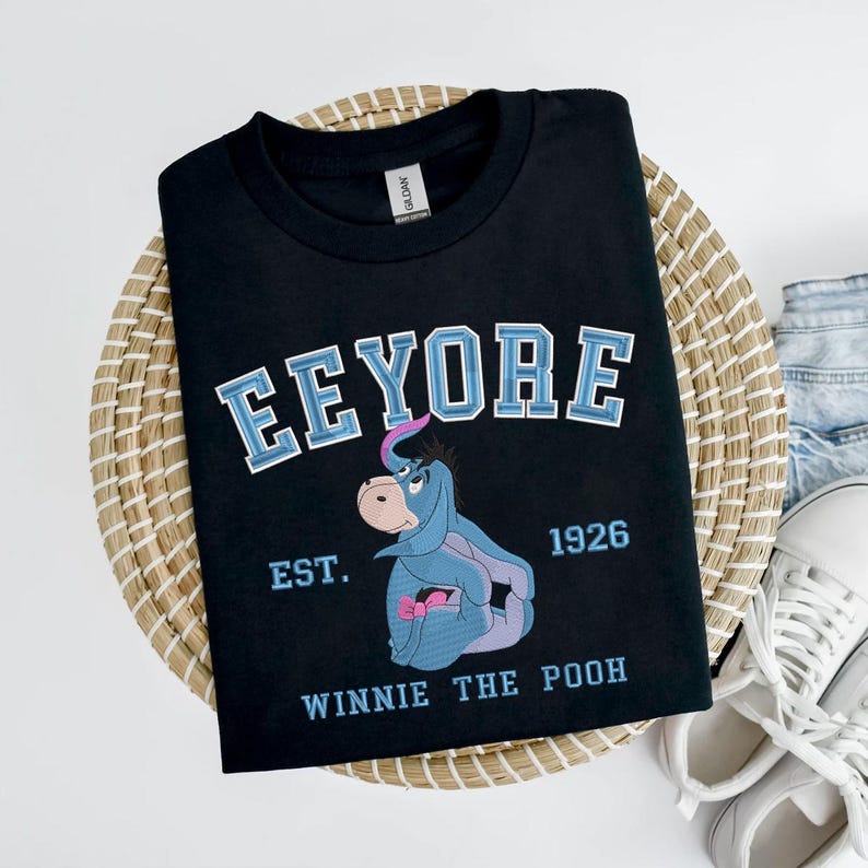 May include: Black t-shirt featuring a graphic of Eeyore from Winnie the Pooh. The design includes the name "Eeyore" in blue and white lettering, the year "1926", and the phrase "Winnie the Pooh". The shirt is displayed on a woven basket.