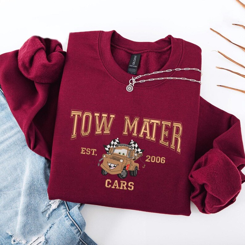 Puede incluir: Sudadera burdeos de cuello redondo con el texto "TOW MATER" y una imagen de dibujos animados de Mater de la pel&iacute;cula Cars. La sudadera tambi&eacute;n tiene el texto "EST. 2006" y "CARS". Un collar plateado est&aacute; drapeado sobre la sudadera.