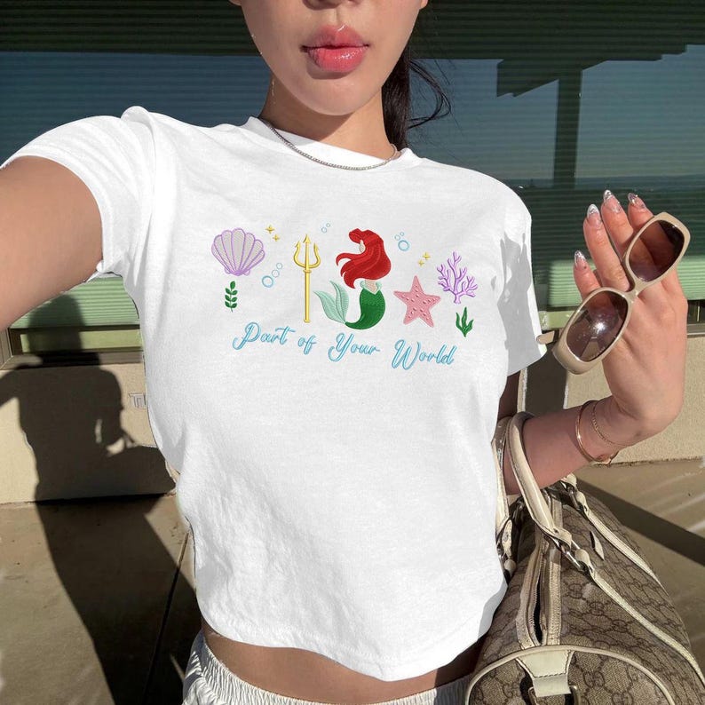 May include: White crop top with a mermaid-themed graphic. The design includes a mermaid, a trident, a seashell, and the text "Part of Your World". The top is a casual, everyday wear item. The text is in a whimsical font.