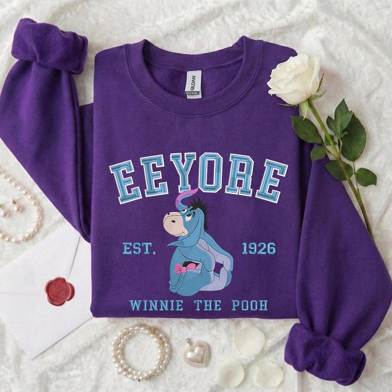 May include: A purple sweatshirt featuring the character Eeyore from Winnie the Pooh. The sweatshirt has the word "Eeyore" in light blue lettering, the text "EST. 1926", and the words "Winnie the Pooh". A white rose and pearl necklace are also in the image.