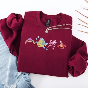 May include: Burgundy crewneck sweatshirt with embroidered cartoon characters, including a tiara, a yellow fish, a pink bow, and a red crab. A silver necklace with a pendant is draped across the sweatshirt. The sweatshirt is folded on top of blue jeans.