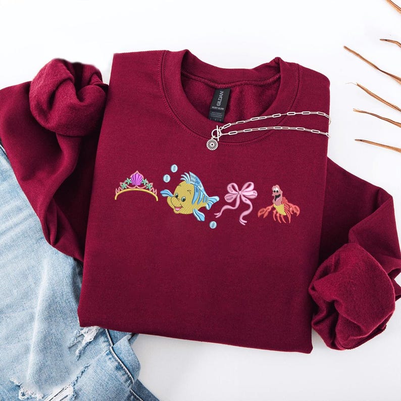 May include: Burgundy crewneck sweatshirt with embroidered cartoon characters, including a tiara, a yellow fish, a pink bow, and a red crab. A silver necklace with a pendant is draped across the sweatshirt. The sweatshirt is folded on top of blue jeans.